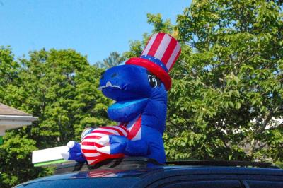 A patriotic t-rex sticks out of a car's sunroof. 