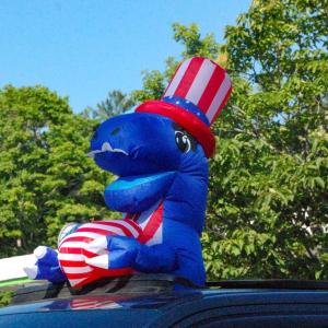 A patriotic t-rex sticks out of a car's sunroof. 
